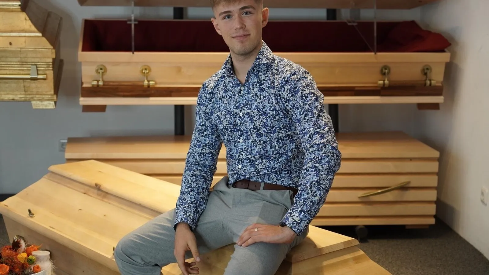 Der angehende Bestatter Luis Bauer sitzt auf einem Holz-Sarg für ein Foto.